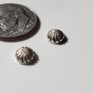 May include: Two small, silver-colored, ribbed bead caps are displayed next to a U.S. dime. The bead caps have a rounded, fluted design, and are likely used in jewelry making or crafting. The dime provides a size reference.