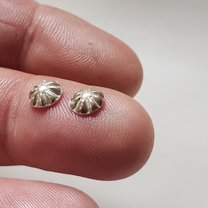May include: Two small, silver-colored, flower-shaped jewelry components. Each component has a detailed, ridged design, resembling a stylized flower or sunburst. The components are shown close-up, highlighting their intricate details and metallic sheen.