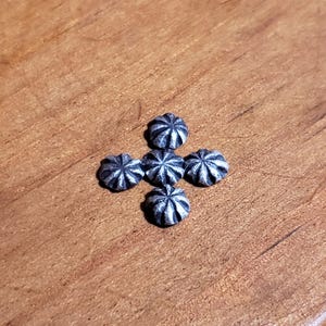 May include: Five small, silver-colored, star-shaped decorative buttons arranged in a cross pattern. The buttons have a detailed, textured design and are set against a warm, brown wooden surface.