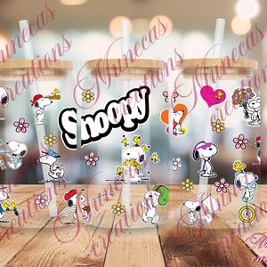May include: Three clear glass tumblers with bamboo lids and straws, decorated with Snoopy and Woodstock stickers, flowers, and hearts. The word "Snoopy" is displayed in a black and white font. The tumblers are on a wooden surface.