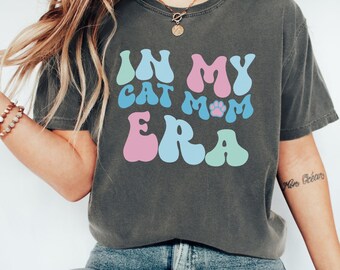 Könnte beinhalten: Ein dunkelgraues T-Shirt mit dem Text "IN MY CAT MOM ERA" in bunten, Retro-Lettern. Der Text enthält einen Pfotenabdruck.