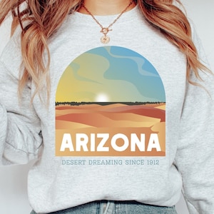 May include: A light gray sweatshirt featuring a desert landscape design with the word "ARIZONA" in white block letters. The design includes a sunset over sand dunes and the text "DESERT DREAMING SINCE 1912".