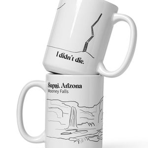 Könnte beinhalten: Zwei weiße Keramikbecher, die übereinander gestapelt sind. Der obere Becher hat eine schwarze Strichzeichnung einer Leiter, die an einer Klippe lehnt, mit dem Text "Ich bin nicht gestorben." Der untere Becher hat eine schwarze Strichzeichnung der Mooney Falls in Supai, Arizona, mit dem Text "Supai, Arizona Mooney Falls."