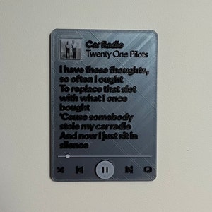 May include: A gray rectangular plaque with the text "Car Radio" by Twenty One Pilots. The plaque features song lyrics and a music player interface with play, skip, and pause buttons. A small photo of the band is at the top.