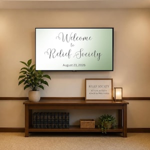 Puede incluir: Un televisor de pantalla plana muestra el texto Welcome to Relief Society August 23, 2026. Debajo, una mesa consola de madera sostiene una planta en maceta, un letrero enmarcado que dice Relief Society Action in Faith Power in Priesthood y una lámpara.