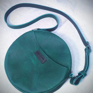 May include: A round, green leather crossbody bag with a black strap and a small, black leather patch with the word "Handmade" on it.