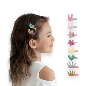 May include: A young girl with brown hair wearing a white top, with several decorative hair clips. The clips are shaped like hearts, stars, crowns, and bunnies, and are decorated with glitter in various colors, including pink, gold, and teal.