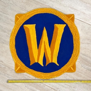 May include: A circular tufted rug featuring a large yellow letter W centered on a deep blue background, surrounded by a thick yellow border. A yellow measuring tape is placed along the bottom, showing a width of approximately 24 inches.