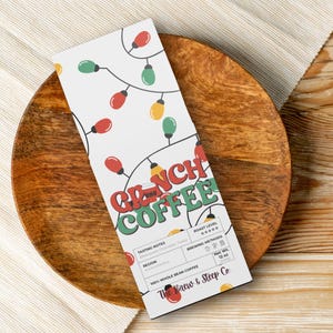 May include: A white coffee bag with the text "Grinch Coffee" in red and green, decorated with colorful Christmas lights. The bag is on a wooden plate, with a beige cloth and wooden surface in the background. The bag is labeled "100% Whole Bean Coffee".