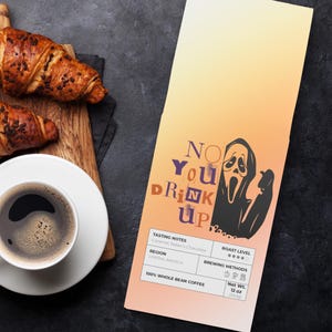 May include: Overhead view of a coffee cup, two chocolate croissants on a wooden board, and a coffee bag. The bag has a gradient background and a ghost graphic with the text "NO YOU DRINK UP". The croissants are golden brown.