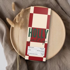 May include: A rectangular coffee box with a red and cream checkerboard pattern. The box has the words "HOLLY jolly" in green and red. The box is on a wooden tray with dried plants and a tan cloth.