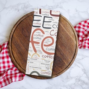May include: A coffee bag featuring the word "coffee" in diverse fonts and shades of brown, beige, and black. It rests on a wooden serving board, accompanied by a red and white checked cloth.