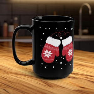 Könnte beinhalten: Schwarze Keramik-Tasse mit einem Design aus zwei roten Fäustlingen mit weißen Schneeflockenmustern und weißem Besatz. Die Tasse hat einen großen Henkel und ist mit weißen Punkten verziert, die fallenden Schnee andeuten. Winterliches Thema.