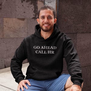 Peut inclure: Un sweat à capuche gris foncé avec le texte blanc "GO AHEAD CALL HR". Le sweat à capuche a une capuche à cordon et une poche avant. La personne sur l'image porte un short bleu.
