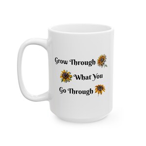 以下が含まれることがあります： 黒い花と黄色のひまわり模様と「Grow Through What You Go Through」という文字が書かれた白いセラミック製のコーヒーカップです。