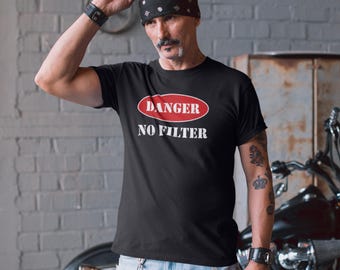 May include: Black t-shirt with the text "DANGER NO FILTER" in a red and white design. The shirt is short-sleeved and worn by a person with a bandana and tattoos. The background includes a brick wall and a motorcycle.