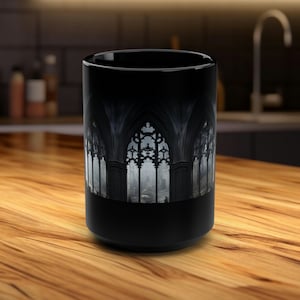 Op de afbeelding: Zwarte keramische koffiemok met een gotisch kathedraalraamontwerp. Het raam is omlijst door stenen bogen en heeft ingewikkelde details. De achtergrond is een donker, mistig stadsbeeld.