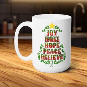 Könnte beinhalten: Weiße Keramik-Kaffeetasse mit einem grünen und roten Weihnachtsbaumdesign. Der Baum ist mit den Worten "Joy", "Noel", "Hope", "Peace" und "Believe" verziert.