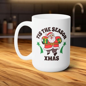 Könnte beinhalten: Weiße Keramiktasse mit einem weihnachtlichen Design. Sie zeigt einen Cartoon-Weihnachtsmann, der Geschenke hält, und den Text "Tis the Season Xmas".