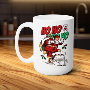 Könnte beinhalten: Weiße Keramik-Kaffeetasse mit einer roten und weißen Illustration von Weihnachtsmann, der einen Schornstein hinunterklettert. Die Tasse trägt den Text "Ho Ho Ho" in roten und grünen Buchstaben.