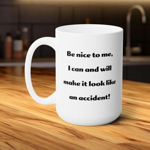 Puede incluir: Taza de cerámica blanca con un asa grande. La taza presenta el texto "Be nice to me, I can and will make it look like an accident!" en negro, centrado en la parte delantera. La taza está sobre una superficie de madera.