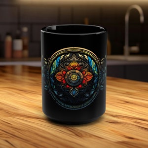 Könnte beinhalten: Schwarze Keramiktasse mit einem farbenfrohen Mandala-Design, das eine Blume mit einem zentralen Auge zeigt. Das Mandala ist von einem kreisförmigen Rahmen mit filigranen Details umgeben.
