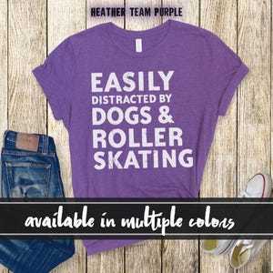 Pode incluir: T-shirt roxo mesclado com o texto branco "EASILY DISTRACTED BY DOGS & ROLLER SKATING". A camisa é exibida com jeans azuis e tênis brancos. O texto "available in multiple colors" também está presente.
