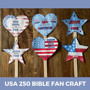 May include: A collection of patriotic paper fan crafts in heart and star shapes. The designs feature the American flag, text that reads "God Bless the USA", "Happy 250th Birthday America!", and religious phrases. Each fan is attached to a wooden stick.