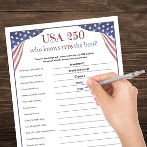 May include: A white paper game sheet with the text "USA 250 who knows 1776 the best?" is being filled out with a pen. The sheet has a blue border with stars and stripes, and includes trivia questions about the year 1776.