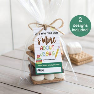 May include: A s'mores treat in a clear cellophane bag, tied with twine, featuring a tag with the text "Let's make this year s'more about Jesus." The tag includes a pencil and apple graphic. A green circle indicates "2 designs included!"