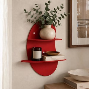 Peut inclure: Une étagère murale rouge avec un panneau arrière ovale et deux niveaux horizontaux. L'étagère supérieure contient un vase en céramique beige avec des branches d'eucalyptus vertes. L'étagère inférieure contient une bougie noire, un bol en bois et deux livres.