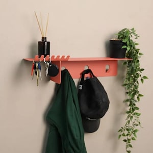 Puede incluir: Estante de pared de color coral con un llavero y ganchos integrados. Una botella difusora negra y una pequeña planta en maceta se encuentran en el estante. Una chaqueta verde y una gorra de béisbol negra cuelgan debajo. Las llaves cuelgan del llavero.