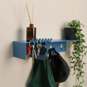 Pu&ograve; includere: Una mensola da parete in metallo blu con una superficie piana e un pannello frontale con diversi ganci integrati. La mensola sostiene una bottiglia in vetro marrone con bastoncini diffusori, un mazzo di chiavi, un tessuto verde e un berretto nero.