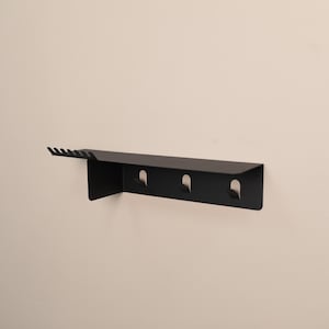 Puede incluir: Un estante de pared de metal negro mate que presenta una superficie superior plana, una fila de seis peque&ntilde;as muescas en el lado izquierdo y tres ganchos integrados que se extienden hacia abajo desde el panel frontal para colgar objetos.