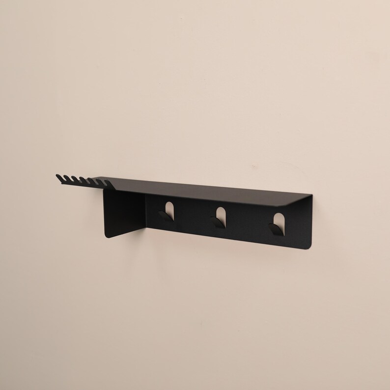 Puede incluir: Un estante de pared de metal negro mate que presenta una superficie superior plana, una fila de seis peque&ntilde;as muescas en el lado izquierdo y tres ganchos integrados que se extienden hacia abajo desde el panel frontal para colgar objetos.