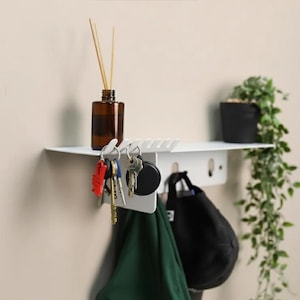 Pu&ograve; includere: Una mensola da parete in metallo bianco con una superficie superiore per riporre oggetti, una fila di ganci per appendere accessori e un pannello posteriore verticale. La mensola sostiene un flacone in vetro marrone, chiavi, un panno verde e un cappellino nero.