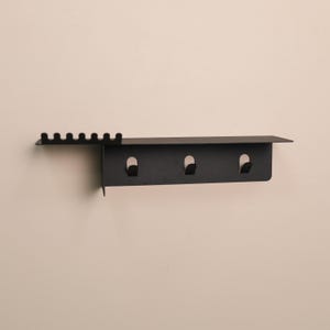 Puede incluir: Un organizador de pared de metal negro mate que cuenta con un estante superior plano, una secci&oacute;n lateral con seis peque&ntilde;as ranuras verticales para colocar objetos y un panel inferior equipado con tres ganchos integrados para colgar diversos art&iacute;culos.