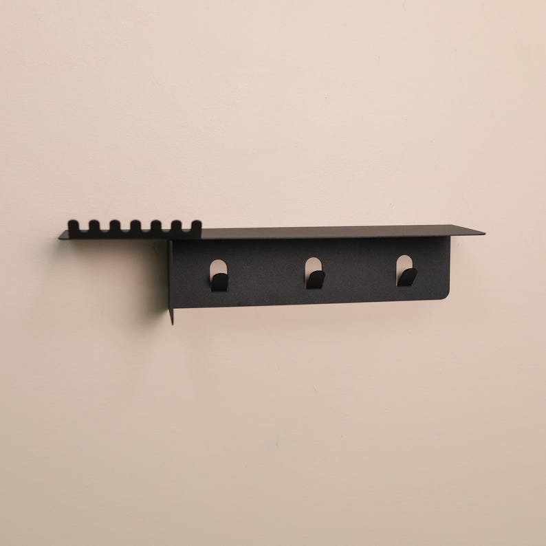 Puede incluir: Un organizador de pared de metal negro mate que cuenta con un estante superior plano, una secci&oacute;n lateral con seis peque&ntilde;as ranuras verticales para colocar objetos y un panel inferior equipado con tres ganchos integrados para colgar diversos art&iacute;culos.