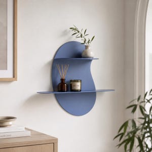 May include: A blue wall-mounted shelf unit featuring a large oval back panel with two horizontal shelves. The top shelf holds a small white vase with a green plant, while the bottom shelf holds a brown glass bottle with reeds and a candle.
