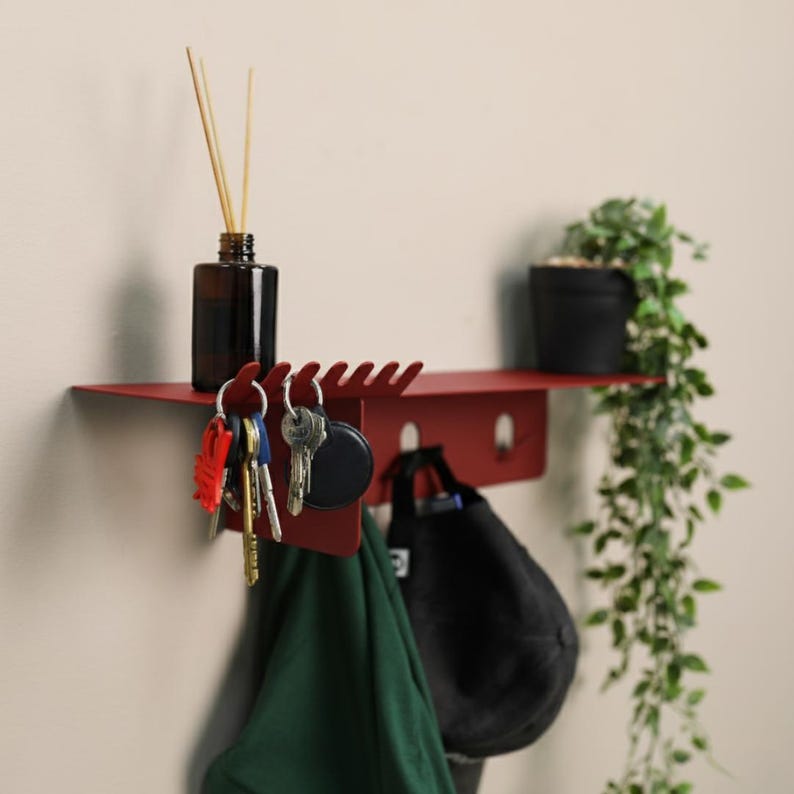 Puede incluir: Un estante de pared de metal rojo oscuro que cuenta con una superficie superior con una fila de ganchos integrados para llaves, un panel vertical con ganchos adicionales, una botella de vidrio oscuro con varillas y una peque&ntilde;a planta verde.