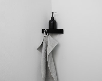 May include: A matte black corner shelf mounted on a white wall, featuring a black pump bottle for liquid soap placed on top. A light gray textured hand towel hangs from a small hook attached to the front edge of the rectangular shelf unit.