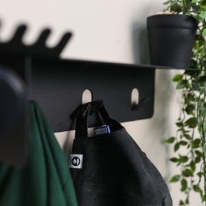 Puede incluir: Un estante de pared de metal negro mate con una repisa superior plana y tres ganchos integrados. El estante presenta un borde ondulado en el lado superior izquierdo. Una gorra de b&eacute;isbol negra cuelga del gancho central y una prenda verde del gancho izquierdo.