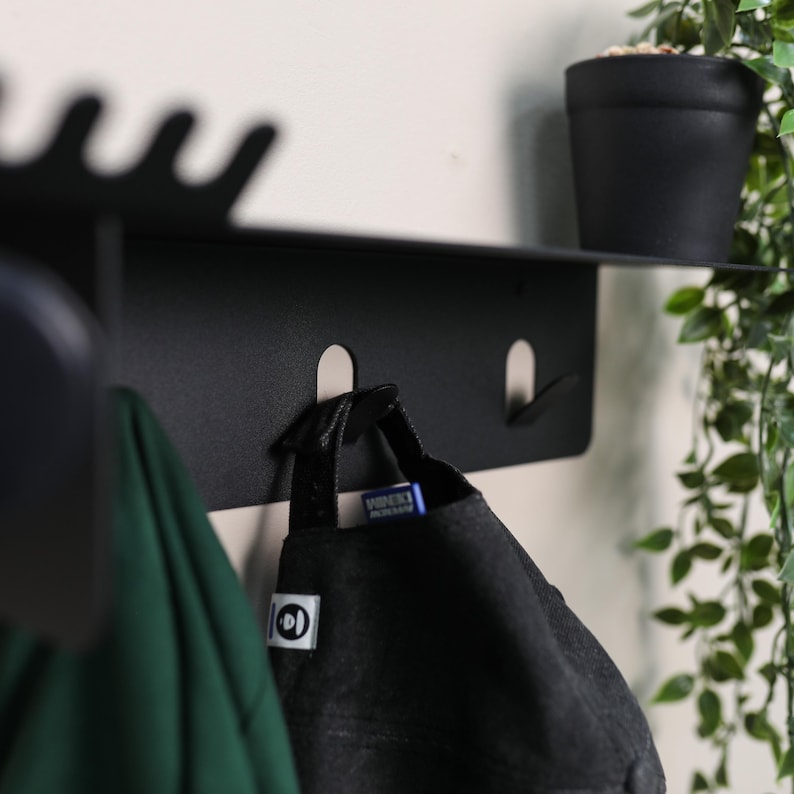 Puede incluir: Un estante de pared de metal negro mate con una repisa superior plana y tres ganchos integrados. El estante presenta un borde ondulado en el lado superior izquierdo. Una gorra de b&eacute;isbol negra cuelga del gancho central y una prenda verde del gancho izquierdo.