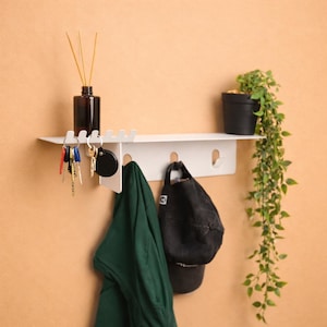 Puede incluir: Un estante de pared blanco que cuenta con una superficie superior con ranuras para colgar llaves, una secci&oacute;n inferior con tres ganchos para prendas o accesorios, un frasco de vidrio marr&oacute;n oscuro con difusores y una peque&ntilde;a planta verde.