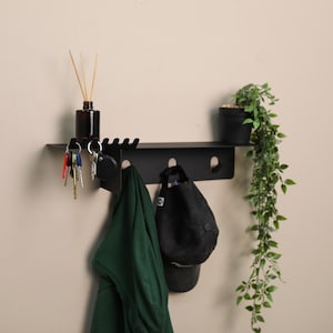 Puede incluir: Un estante de pared de metal negro mate con una superficie superior plana, una fila de ganchos integrados para colgar objetos y una serie de ranuras en el lado izquierdo. El estante sostiene una peque&ntilde;a botella de vidrio marr&oacute;n, una prenda verde y una gorra.