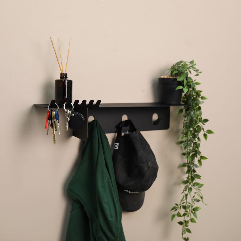 Puede incluir: Un estante de pared de metal negro mate con una superficie superior plana, una fila de ganchos integrados para colgar objetos y una serie de ranuras en el lado izquierdo. El estante sostiene una peque&ntilde;a botella de vidrio marr&oacute;n, una prenda verde y una gorra.