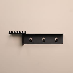 Puede incluir: Un organizador de pared de metal negro mate que cuenta con un estante superior plano, una secci&oacute;n lateral con seis peque&ntilde;as muescas para colgar objetos y un panel vertical debajo del estante equipado con tres ganchos integrados para almacenamiento.