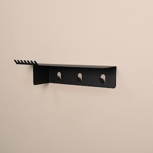 Puede incluir: Un estante de pared negro mate que presenta una superficie superior plana con una serie de seis peque&ntilde;as muescas a lo largo del borde izquierdo y tres ganchos integrados que se extienden hacia abajo desde el panel horizontal principal.