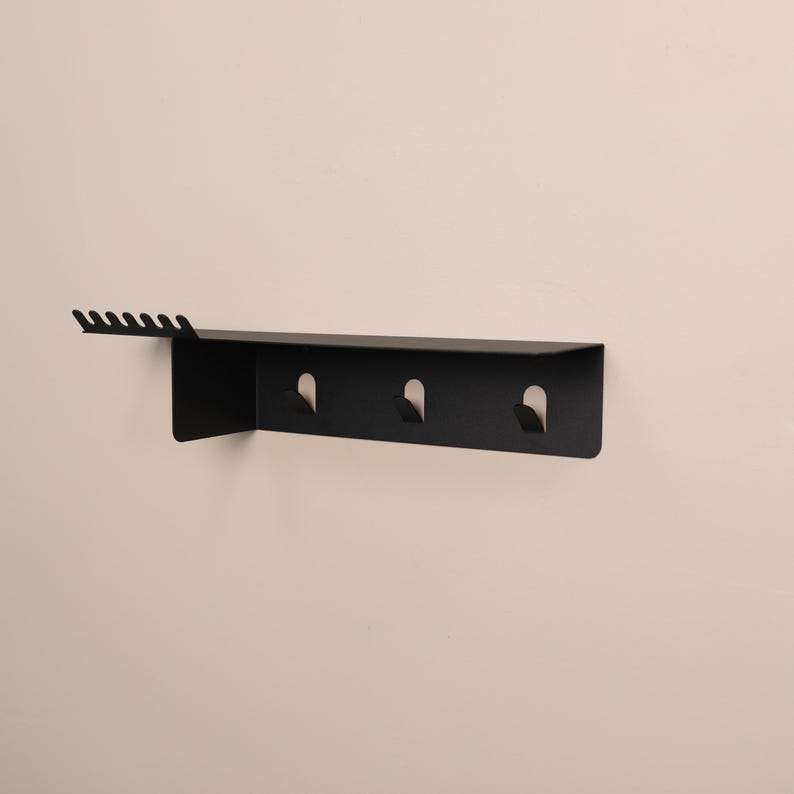 Puede incluir: Un estante de pared negro mate que presenta una superficie superior plana con una serie de seis peque&ntilde;as muescas a lo largo del borde izquierdo y tres ganchos integrados que se extienden hacia abajo desde el panel horizontal principal.
