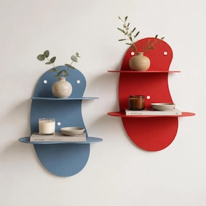 Op de afbeelding: Twee wandplanken met organische, boonvormige achterwanden in blauw en rood. Elke plank heeft twee horizontale oppervlakken met decoratieve items, waaronder keramische vazen, kleine schaaltjes, kaarsen en boeken met de titels Kinfolk en Bau Mangel.